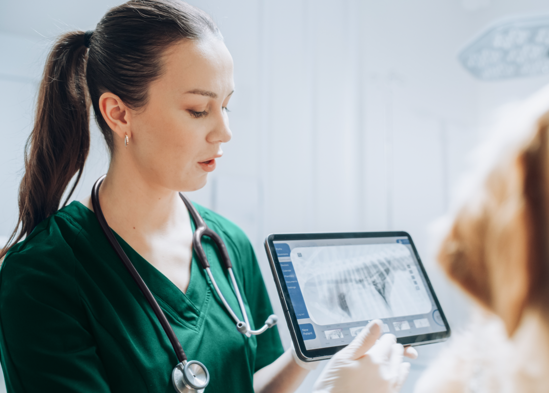 Une vétérinaire, portant un stéthoscope et une blouse teal ANTECH™, présentant des images d'une radiographie sur une tablette, avec le visage flou d'un chien visible au premier plan